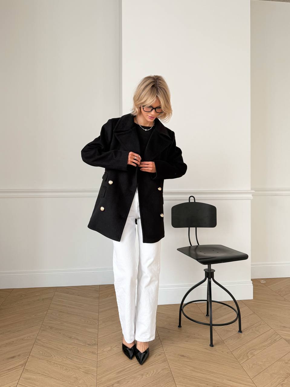 Person wearing a black coat and white pants standing in a room with a black chair.