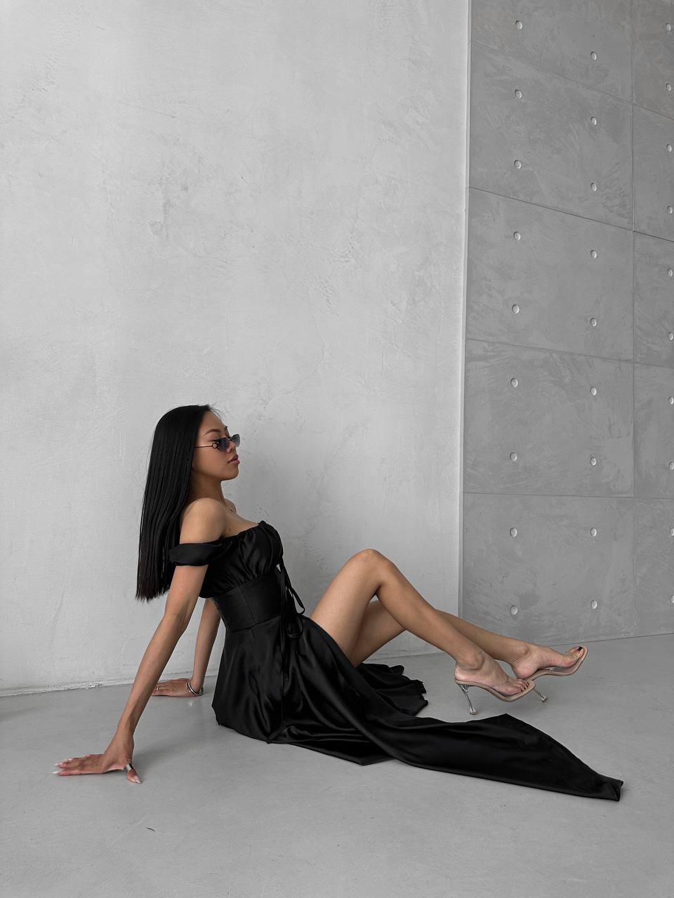 Woman in a black off-shoulder dress sitting against a gray concrete wall.
