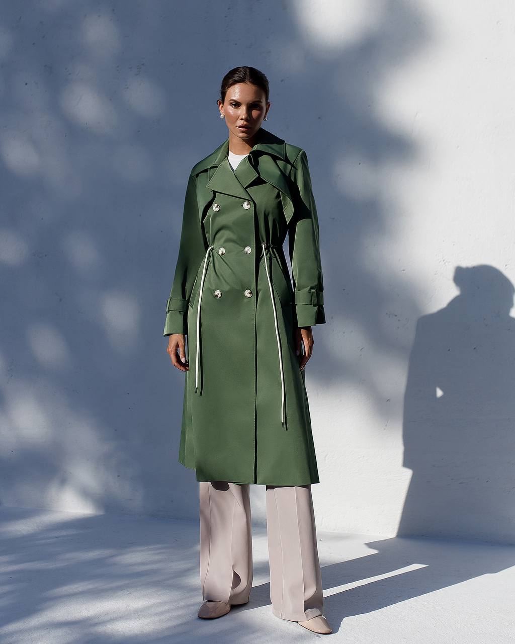 Person wearing a green trench coat standing against a white wall with shadows.