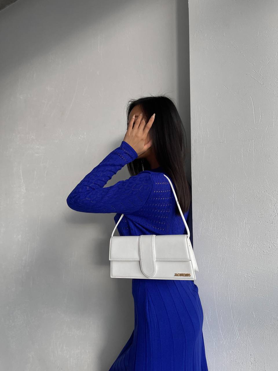 Woman in a blue dress holding a white handbag against a gray wall
