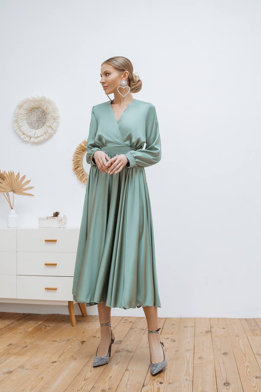 Woman wearing a green dress standing in a room with a white wall and wooden floor.