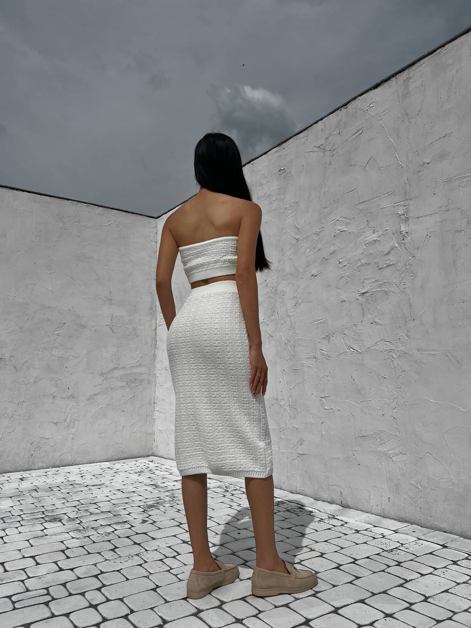 Woman in a white strapless top and skirt standing in a minimalist outdoor setting with gray walls and cobbled floor.