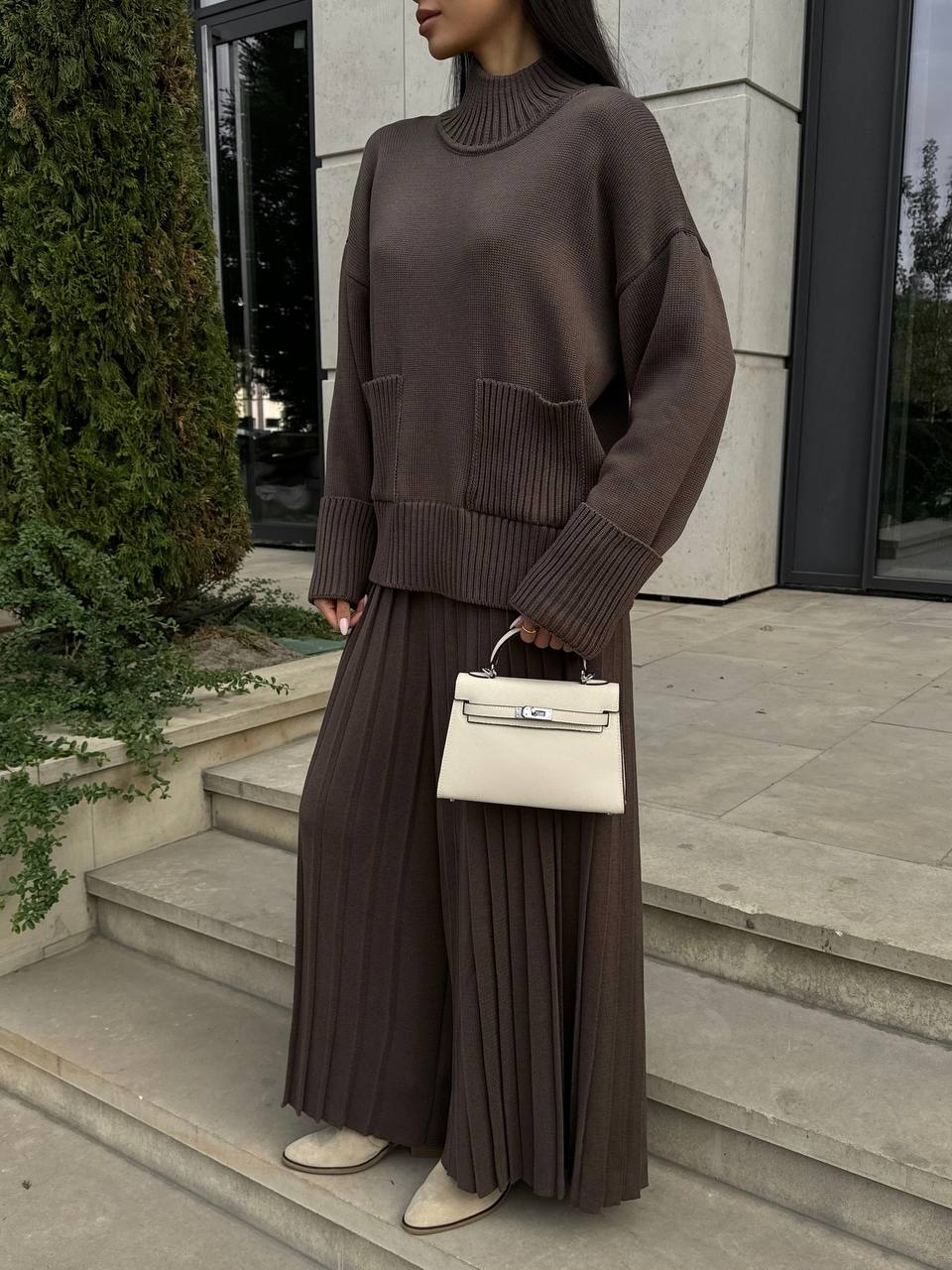 Person wearing a brown sweater and pleated skirt holding a white handbag on steps.