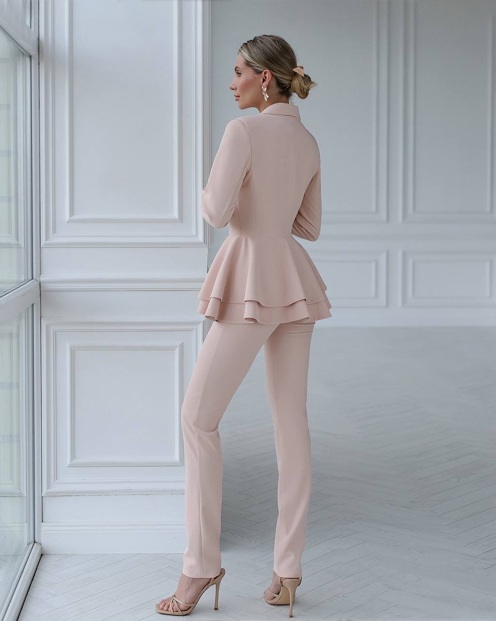 Woman wearing a light pink peplum jacket and pants in a room with white paneled walls.