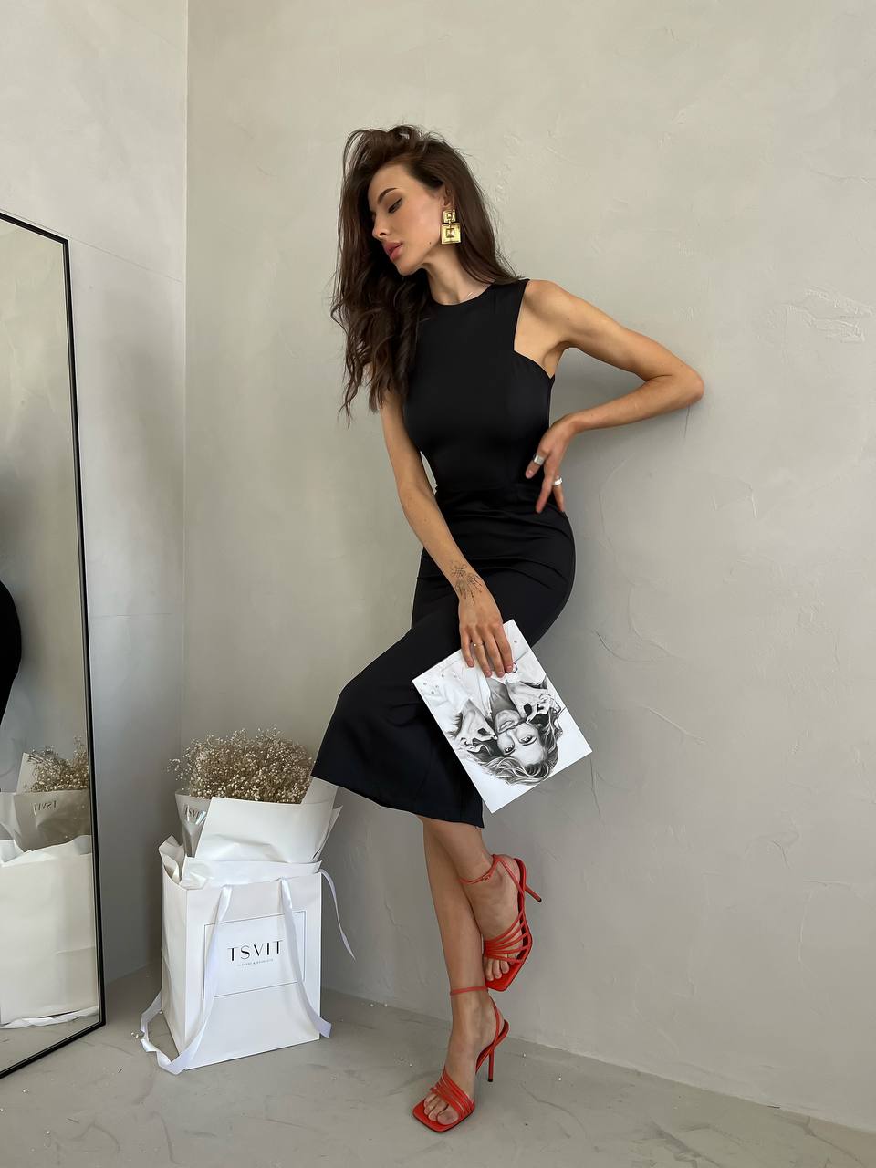 Woman in a black dress with red shoes holding a magazine against a neutral wall.