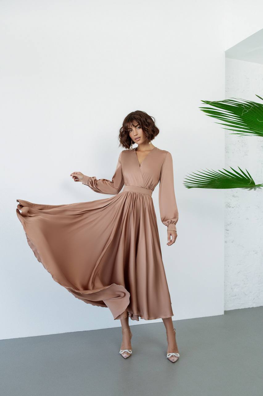 Woman wearing a flowing beige dress against a white background with green plants.