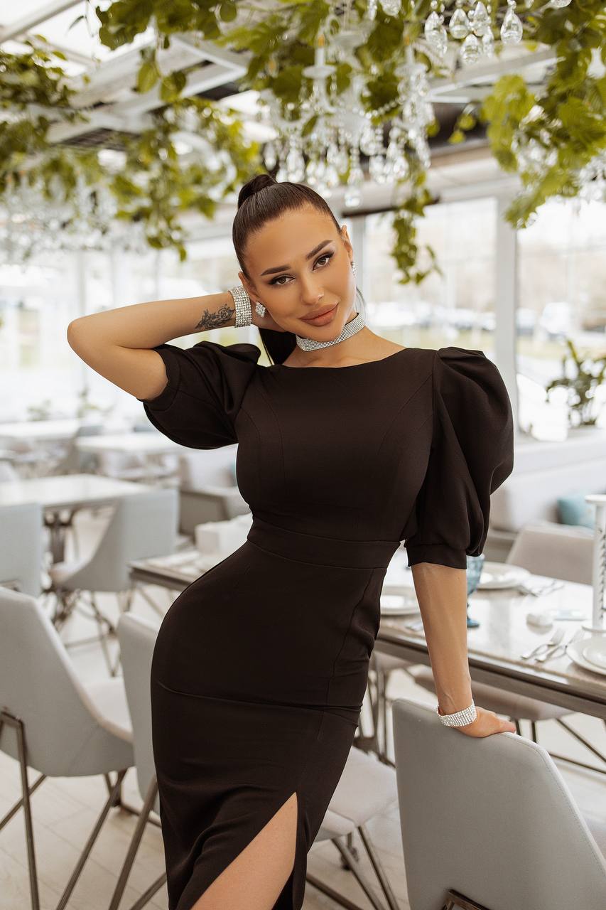 Woman in a black dress posing in an indoor setting with tables and chairs.