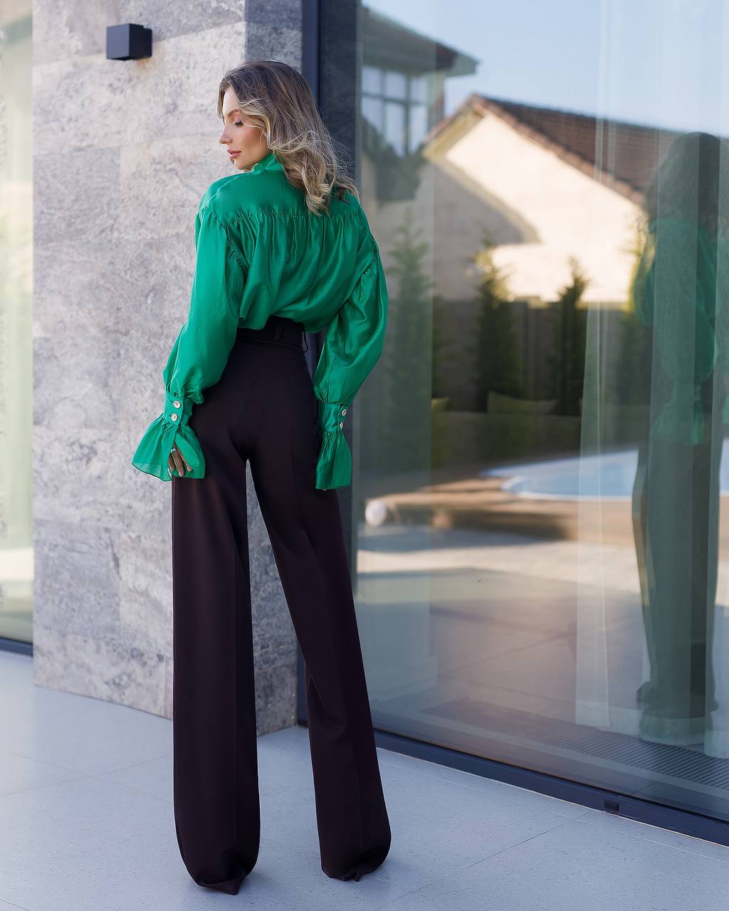 Woman wearing a green blouse and black pants standing in front of a glass door.