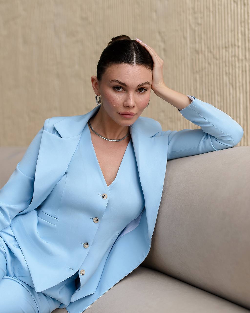 Woman in a light blue suit sitting on a beige couch against a beige wall.