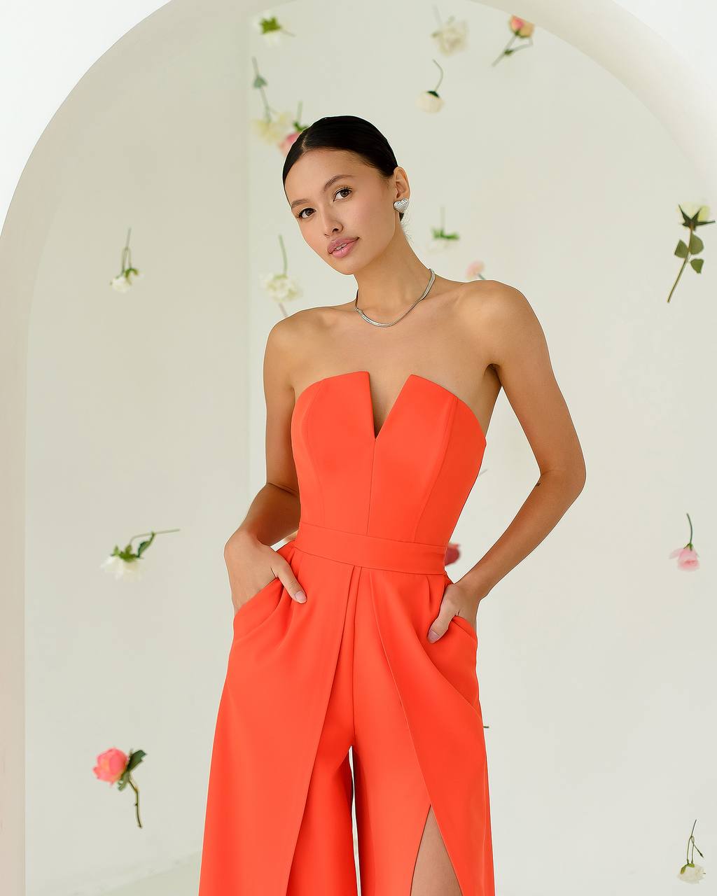 Woman wearing a bright orange strapless jumpsuit with floral decorations in the background
