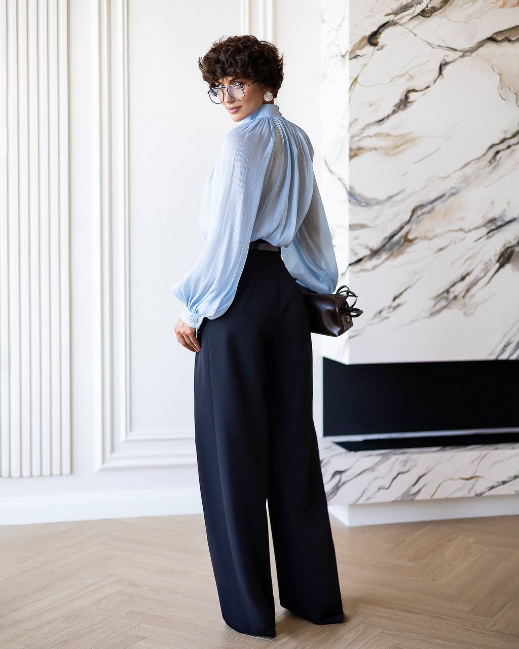 Person wearing a light blue blouse and black pants in an elegant interior setting.