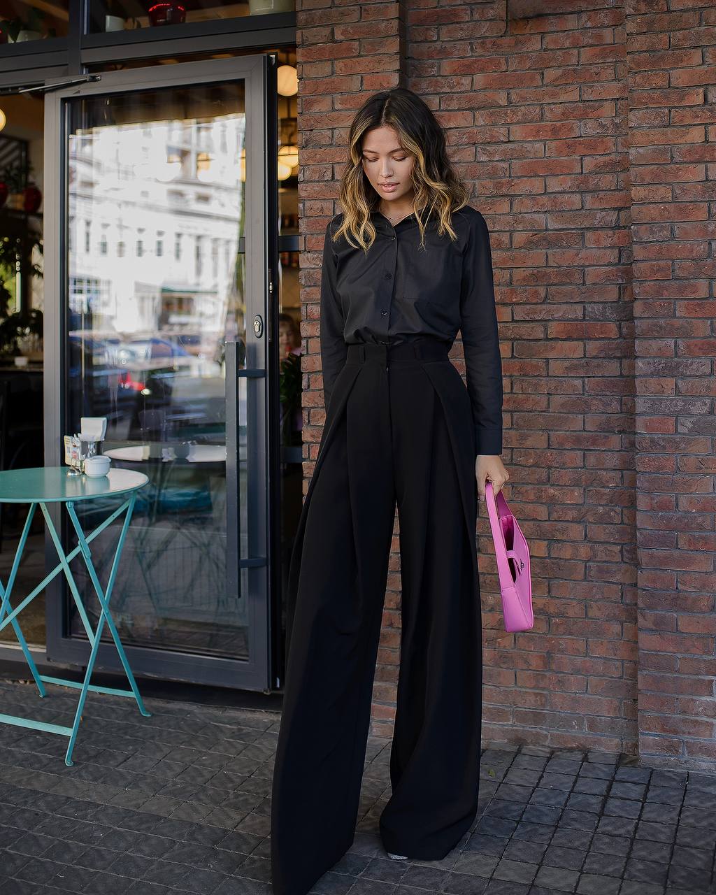 Woman in black outfit holding a pink clutch against a brick wall.