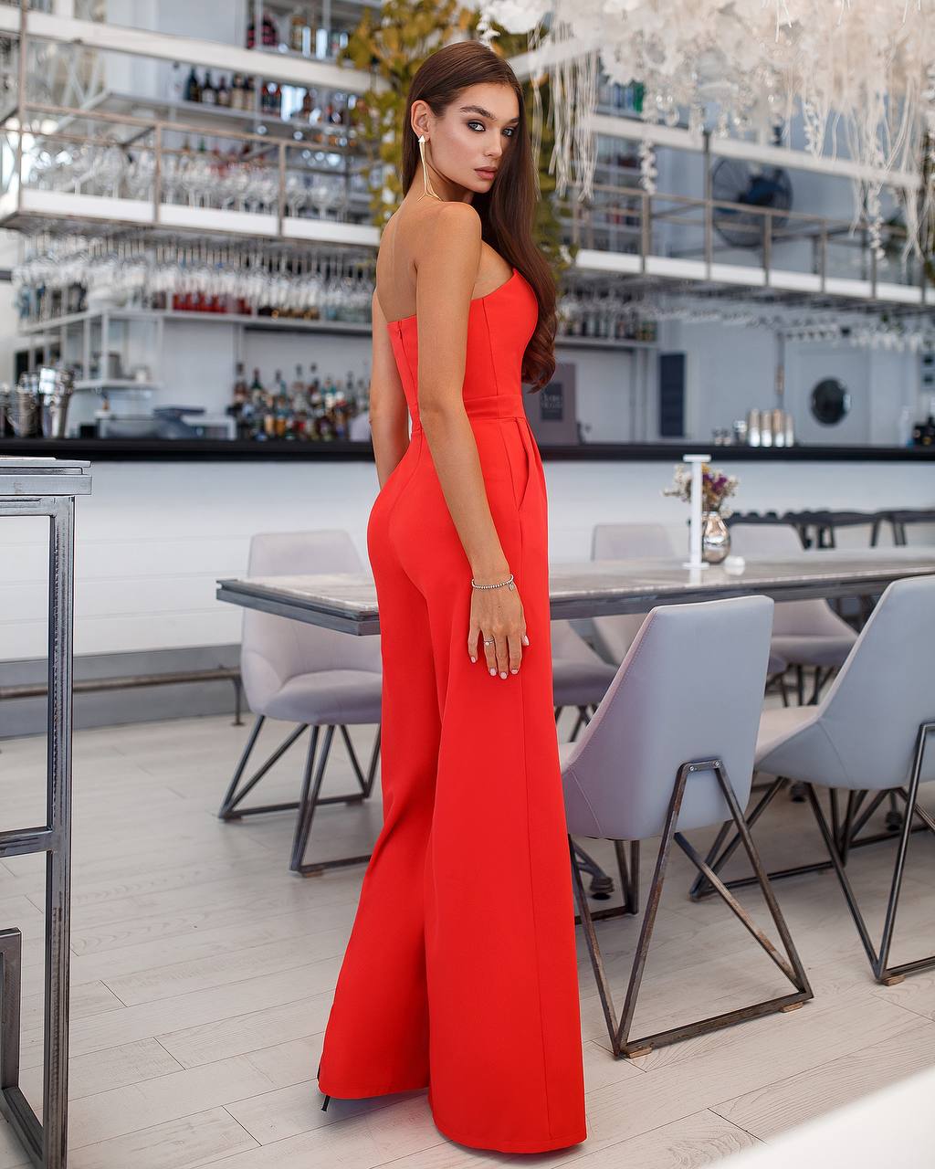 Woman in a red jumpsuit standing in an elegant indoor setting with tables and chairs.