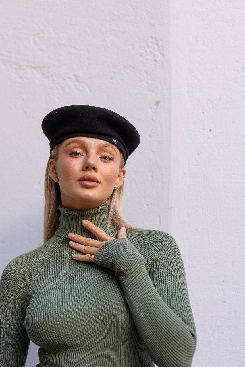Woman wearing a green ribbed turtleneck sweater and black beret against a white wall.