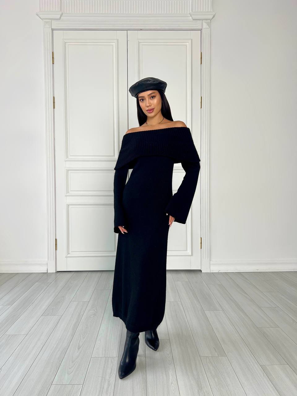 Woman wearing a black off-shoulder dress and beret standing in a room with white walls and wooden floor.