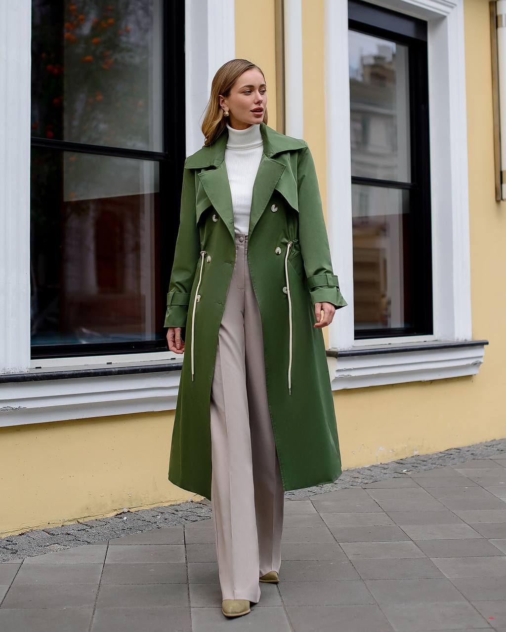 Woman wearing a green coat standing in front of a yellow building with white windows.