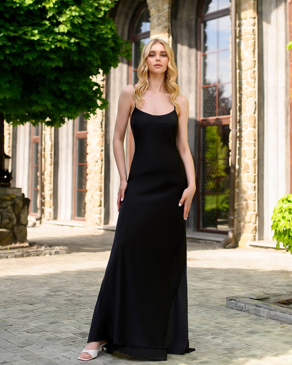 Woman in a black dress standing outdoors with stone building and greenery in the background