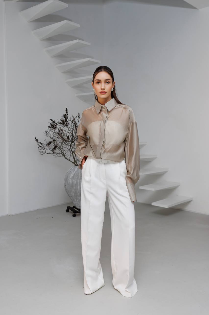Woman in a beige blouse and white pants standing in a minimalistic room with a white staircase.