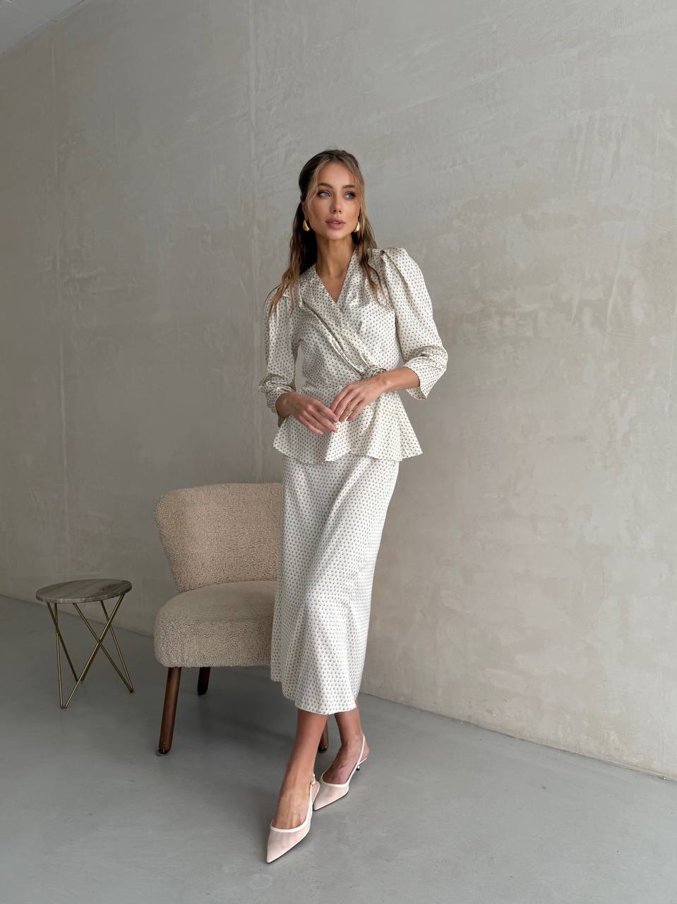 Woman in a white patterned outfit standing in a minimalistic room with a chair and small table.