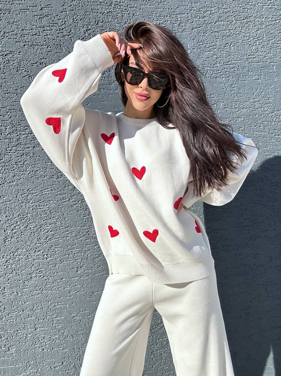 Person wearing a white sweater with red heart patterns against a gray wall.