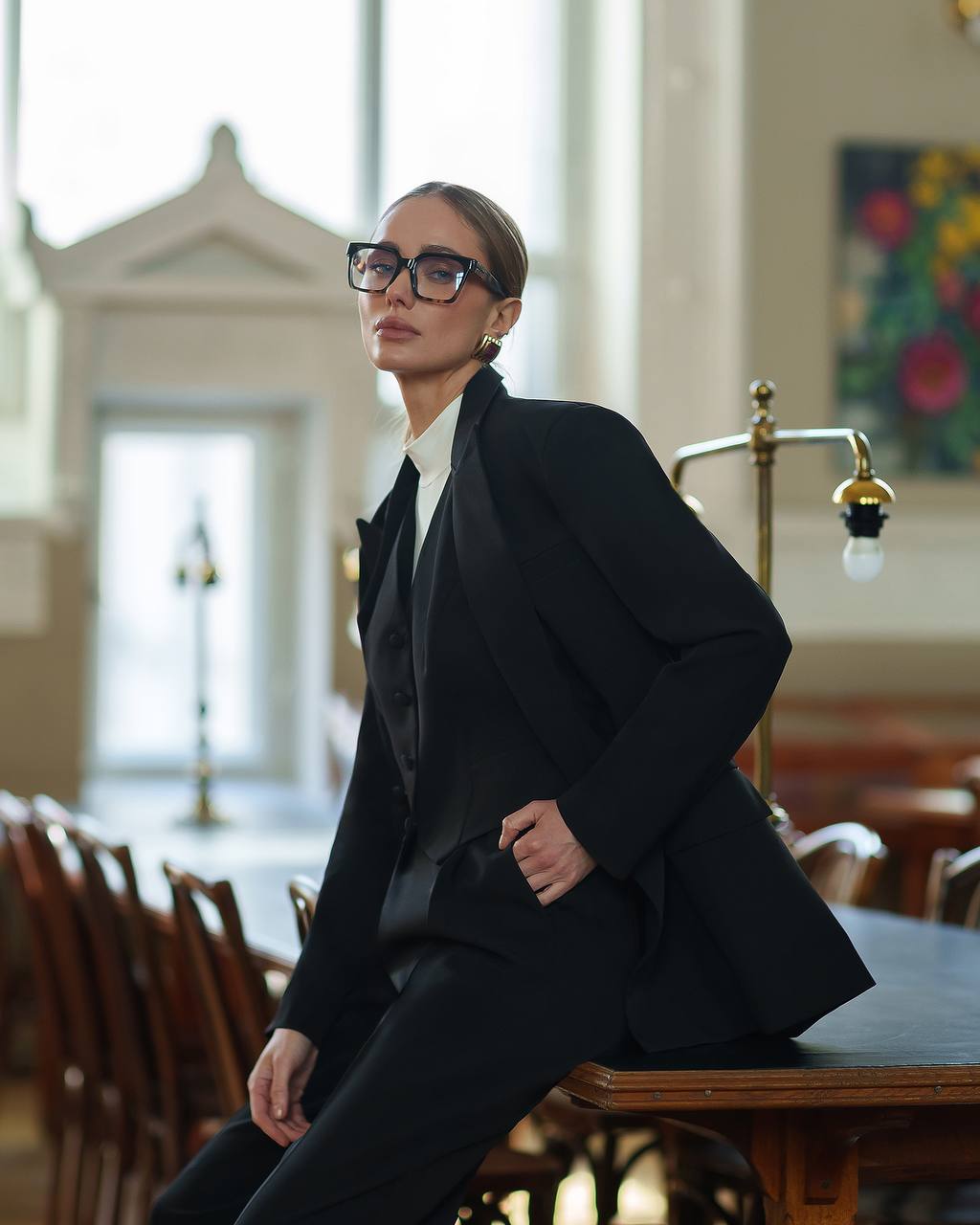 Woman in a black suit sitting in a room with wooden furniture and large windows.