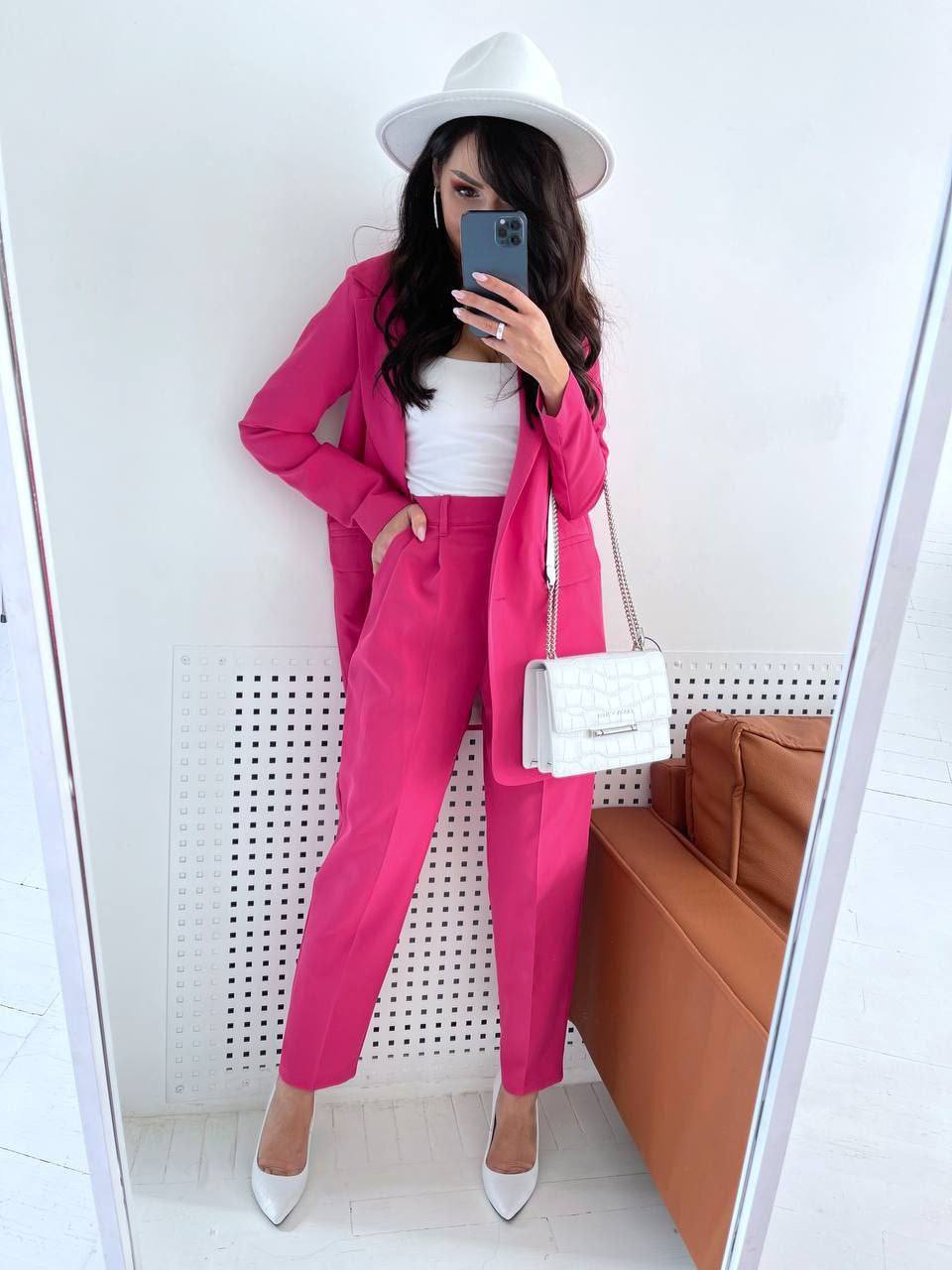 Person wearing a bright pink suit with a white hat and bag, standing in front of a mirror.