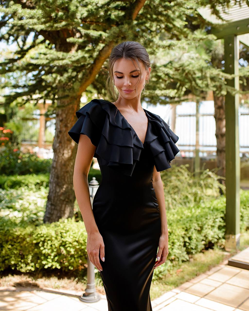 Woman wearing a black ruffled dress in an outdoor setting with greenery.