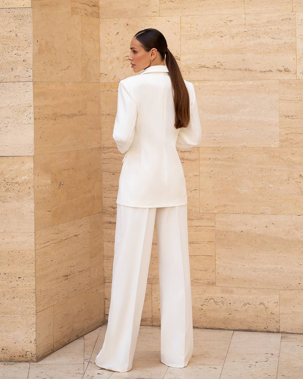 Woman in a white suit standing against a beige stone wall