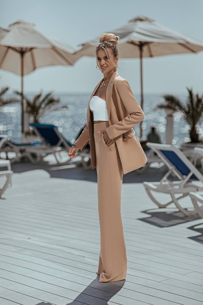 Woman in a beige suit standing on a wooden deck with beach chairs and umbrellas in the background.