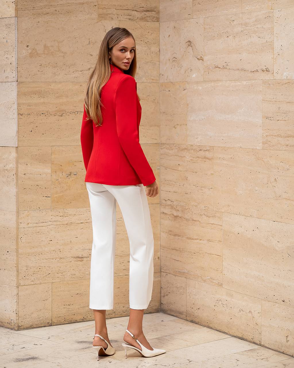 Woman wearing a red blazer and white pants against a beige stone wall.