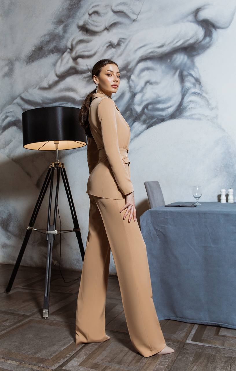 Woman in a beige suit standing in a room with a marble wall and lamp.