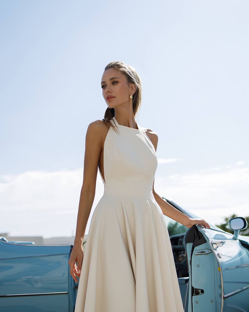 Woman in a white dress standing next to a vintage car with a clear sky background