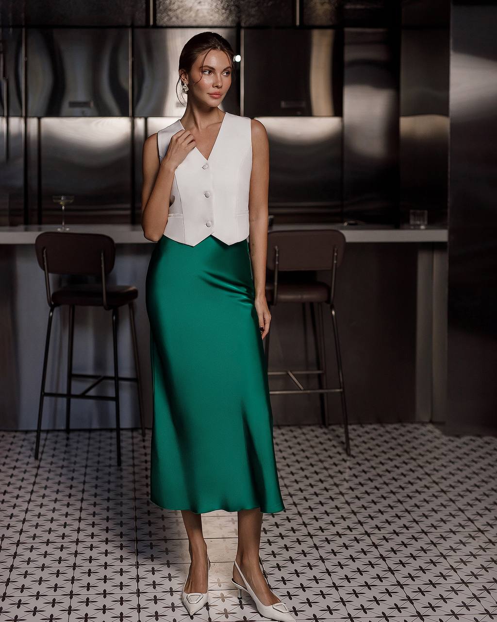 Woman in a white sleeveless top and green skirt standing in a stylish interior setting.