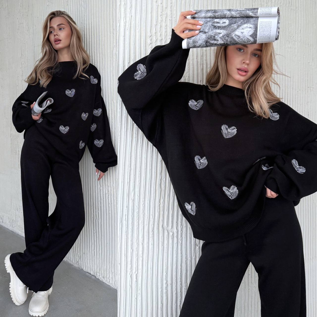 Woman wearing a black sweater with heart patterns against a white textured wall.