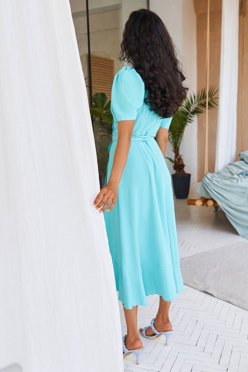 Woman in a turquoise dress standing in a room with white walls and a plant.