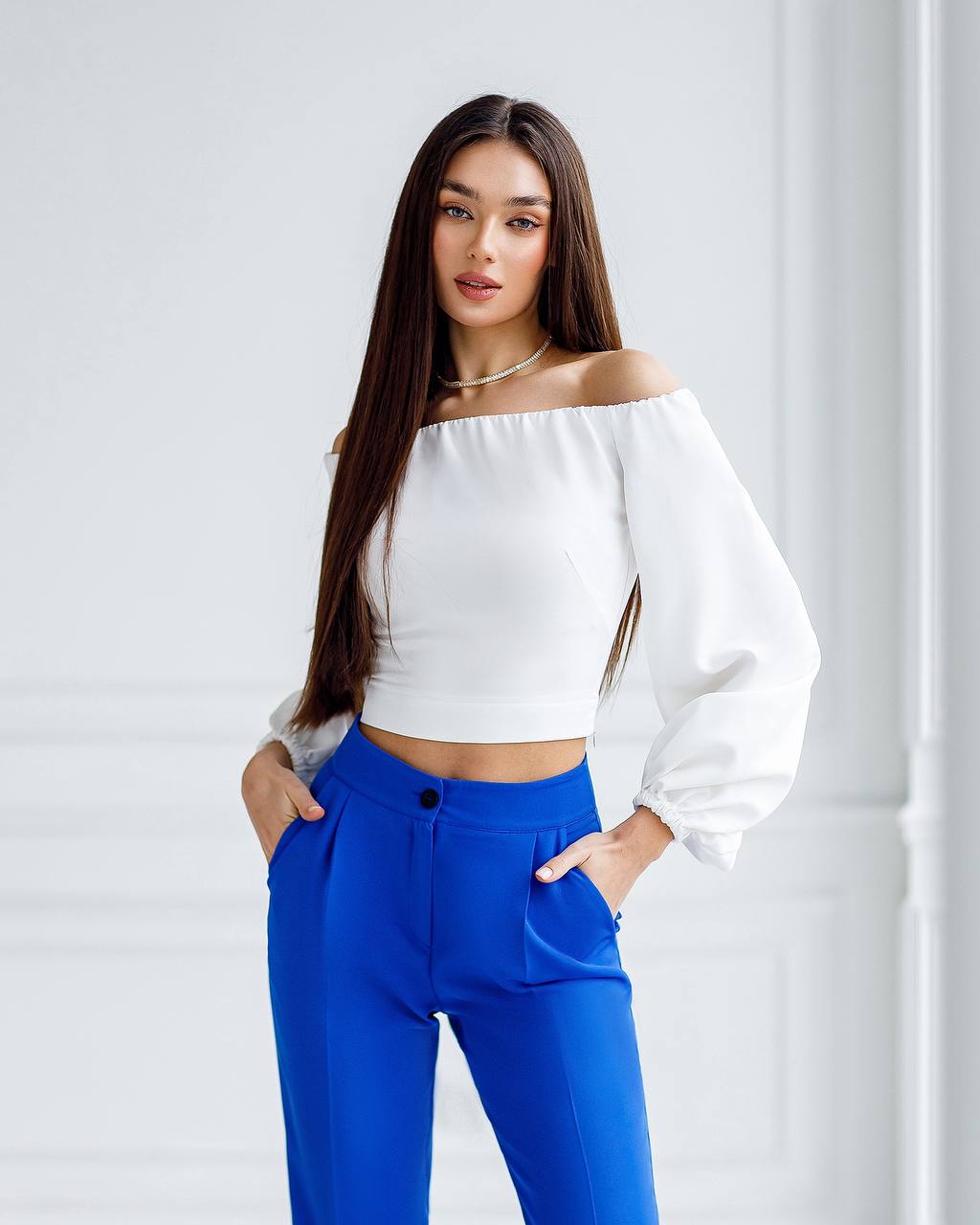 Woman wearing a white off-shoulder top and blue pants against a light gray background