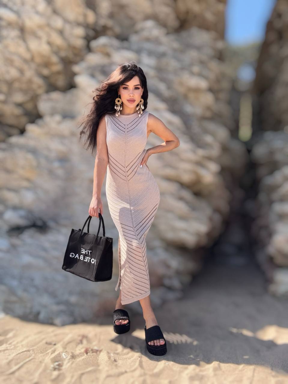 Woman in a sleeveless dress holding a black bag against a rocky background