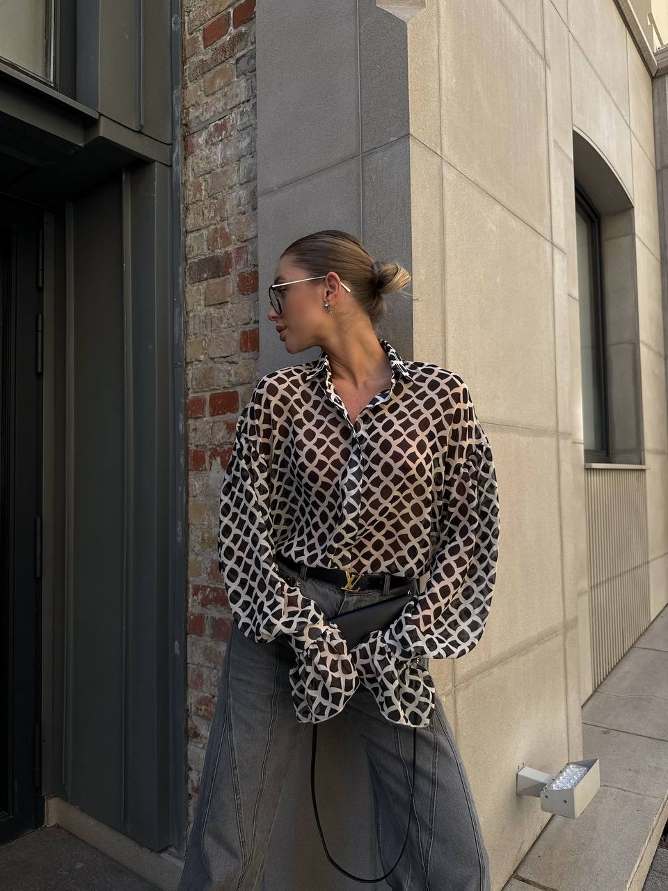 Person wearing a patterned blouse and sunglasses leaning against a building.