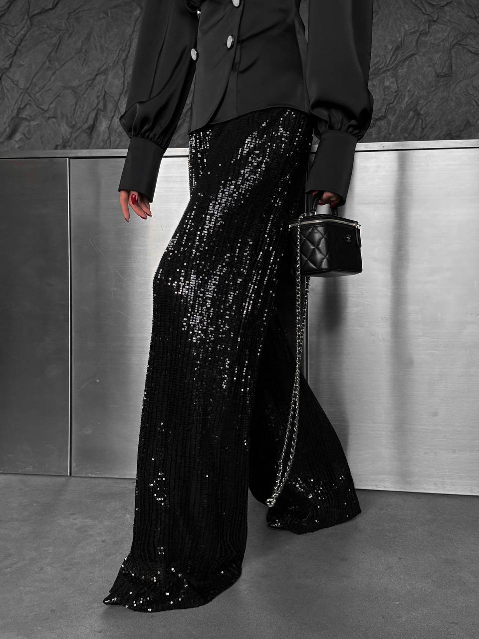Person wearing black sequin pants and a black jacket with a small black handbag against a metallic wall.