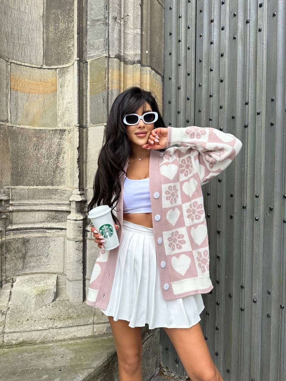 Woman in a pink patterned cardigan and white skirt holding a Starbucks cup against a textured wall.