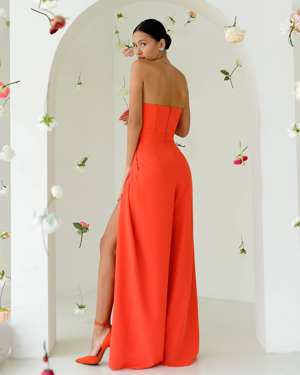 Woman wearing a bright orange strapless jumpsuit with a high slit, standing against a white background with floral decorations.