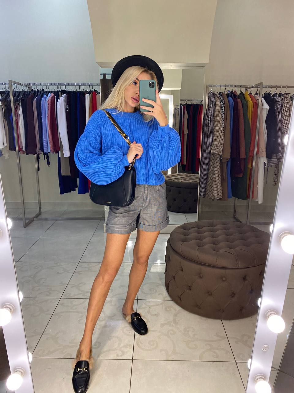 Person taking a mirror selfie in a clothing store wearing a blue sweater and gray shorts.