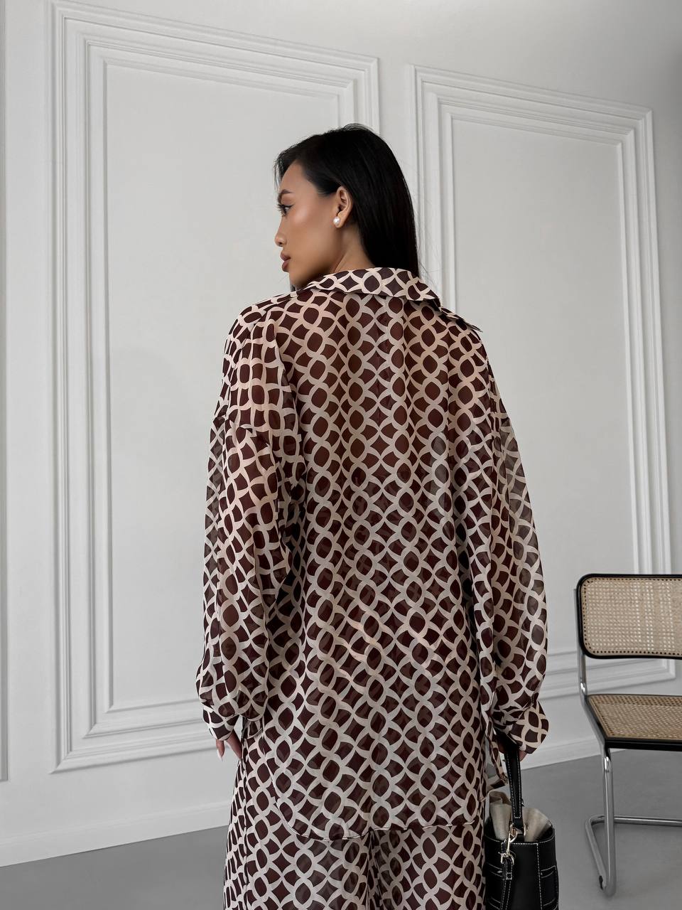Woman wearing a patterned coat in a room with white walls and a chair.
