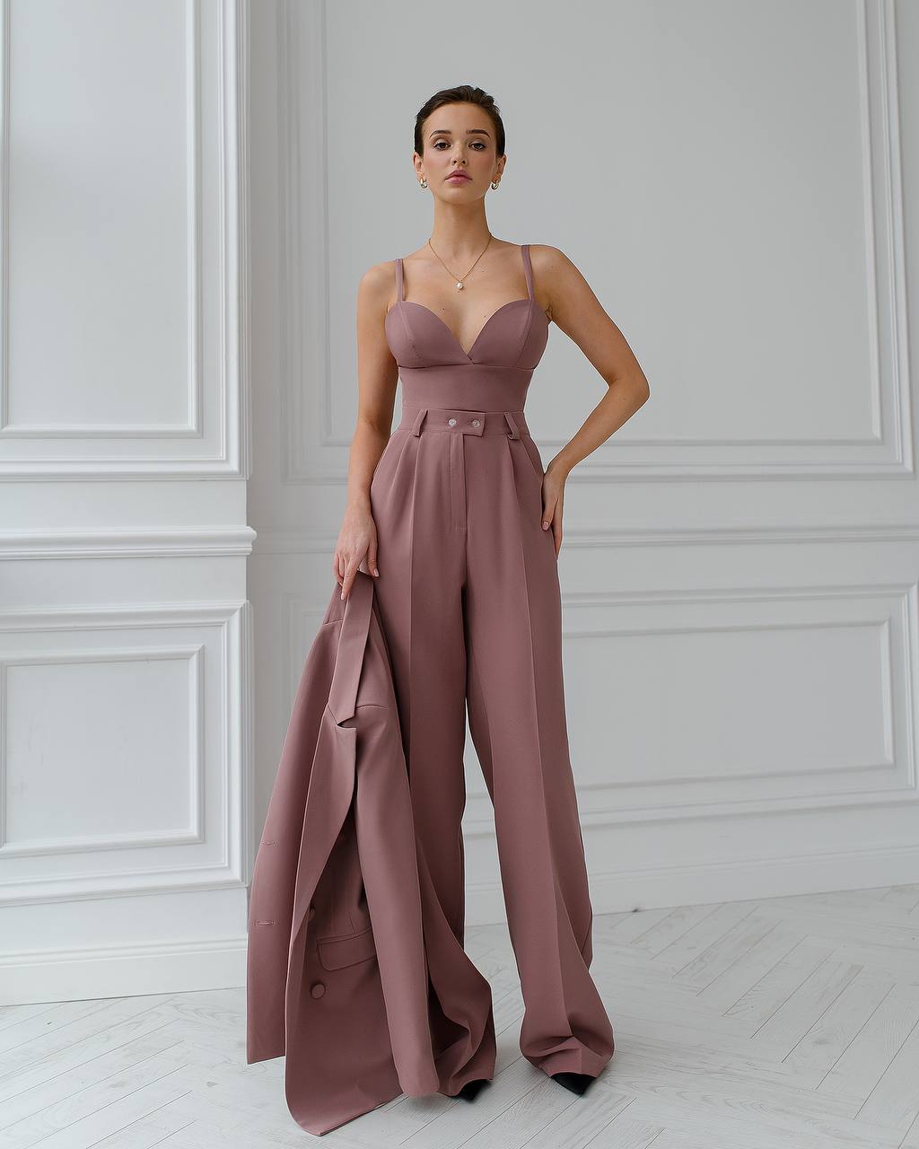 Woman wearing a mauve jumpsuit standing against a white paneled wall.
