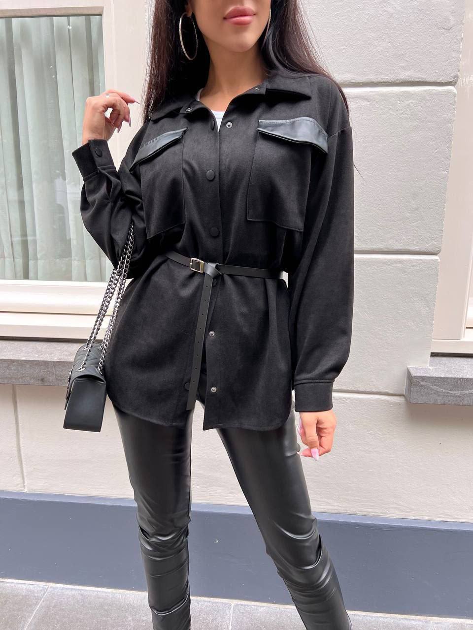 Person wearing a black outfit with leather pants standing against a light-colored wall.