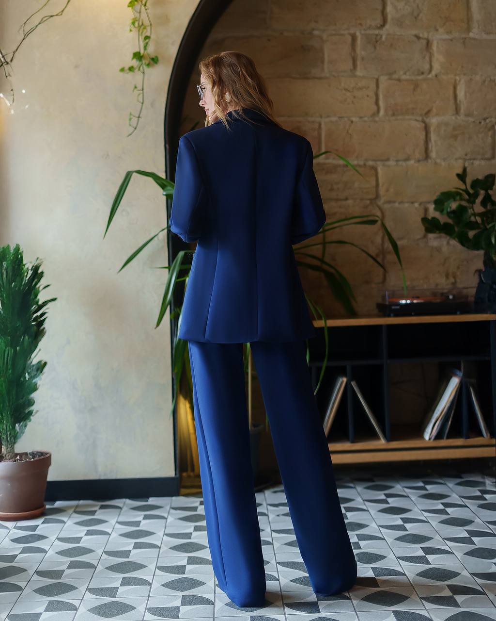 Woman in a blue suit standing in a room with plants and a brick wall.