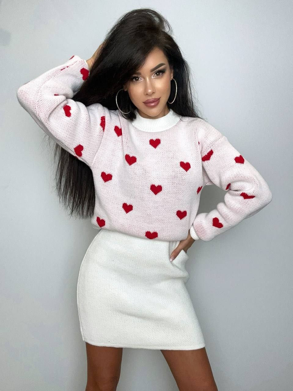 Woman wearing a white sweater with red heart patterns and a white skirt against a plain background