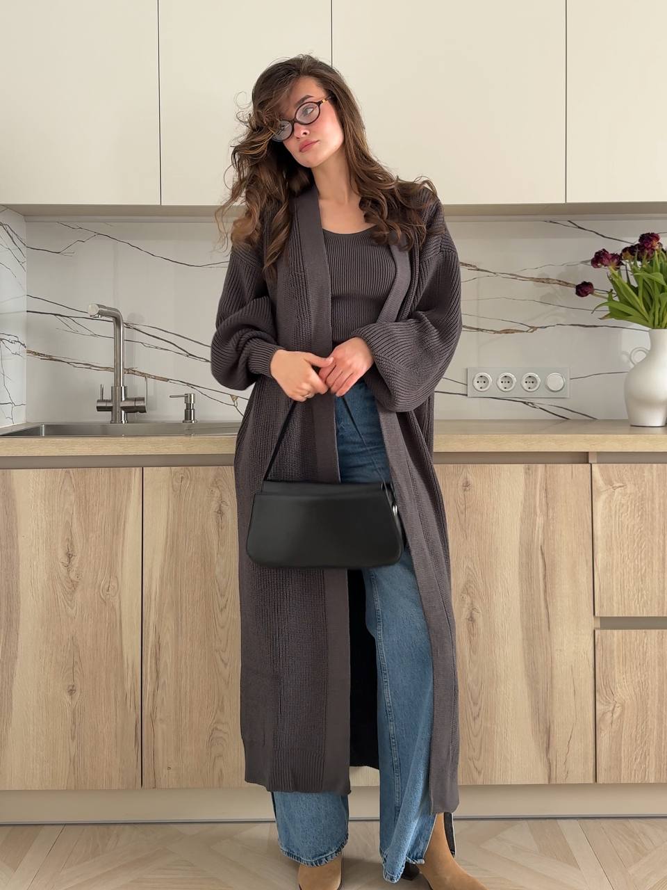 Woman in a long dark gray cardigan and jeans standing in a kitchen.