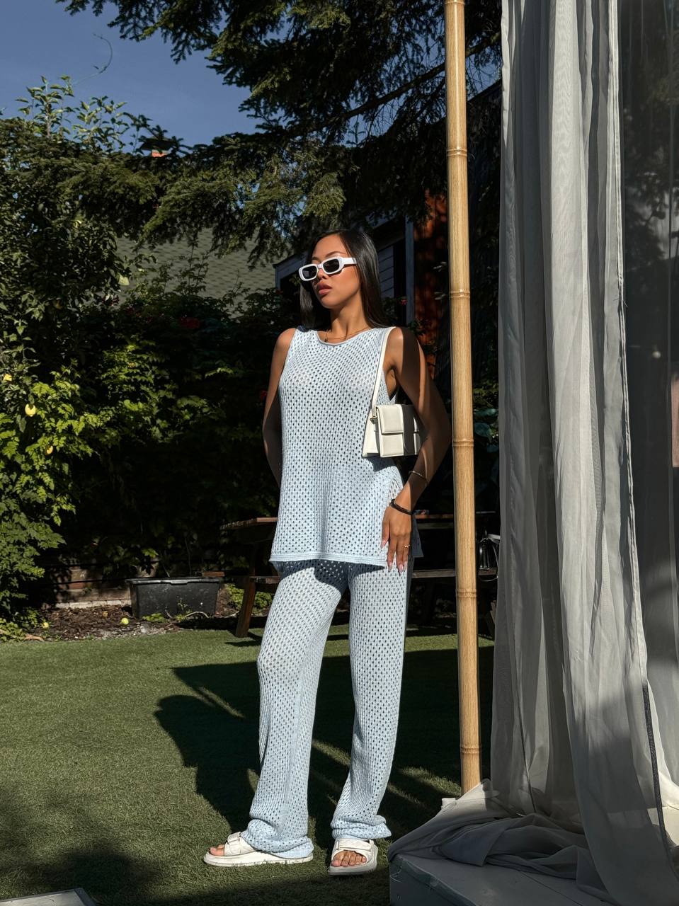 Woman in light blue polka dot outfit standing outdoors with greenery and a bamboo pole in the background.