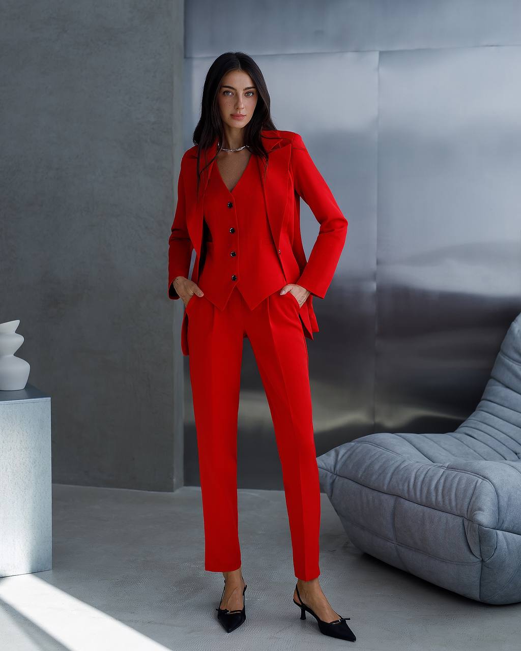 Woman in a red suit standing in a modern interior setting with a gray wall and gray sofa.